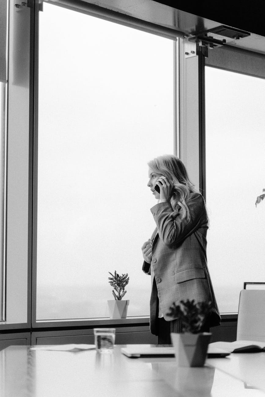 grayscale photo of woman in long sleeve shirt standing beside window